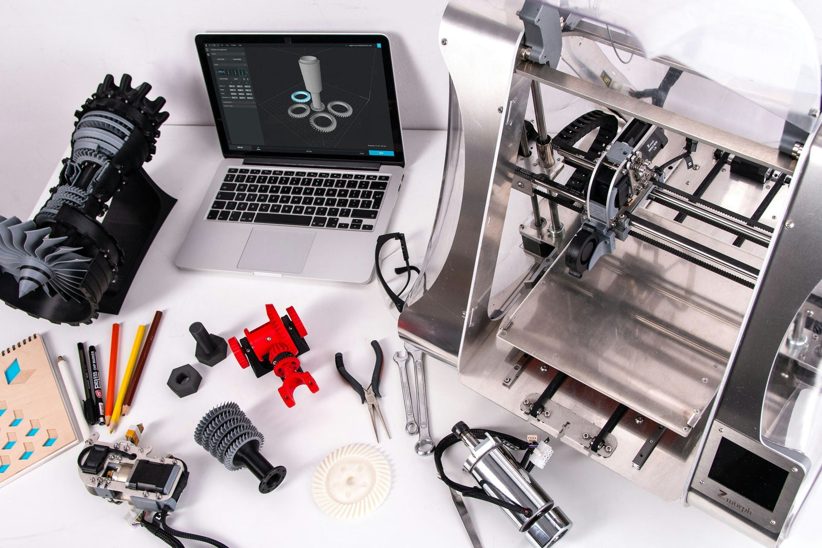 Werkplek met een 3D-printer, laptop met 3D-ontwerpsoftware, gereedschap en verschillende geprinte onderdelen in diverse kleuren op een witte tafel.