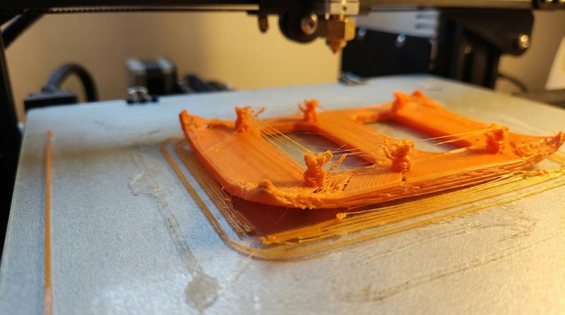 Close-up van 3D printerbed met mislukte print met kromgetrokken hoeken en dunne draden terwijl de nozzle erboven hangt tijdens het printen