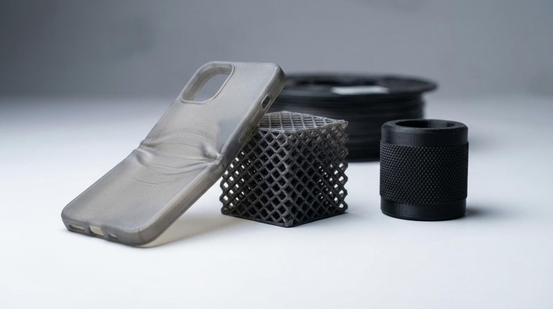Drie TPU 3D prints met verschillende hardheid op grijze ondergrond: flexibele hoes, lattice kubus en cilindergreep met spoel op achtergrond